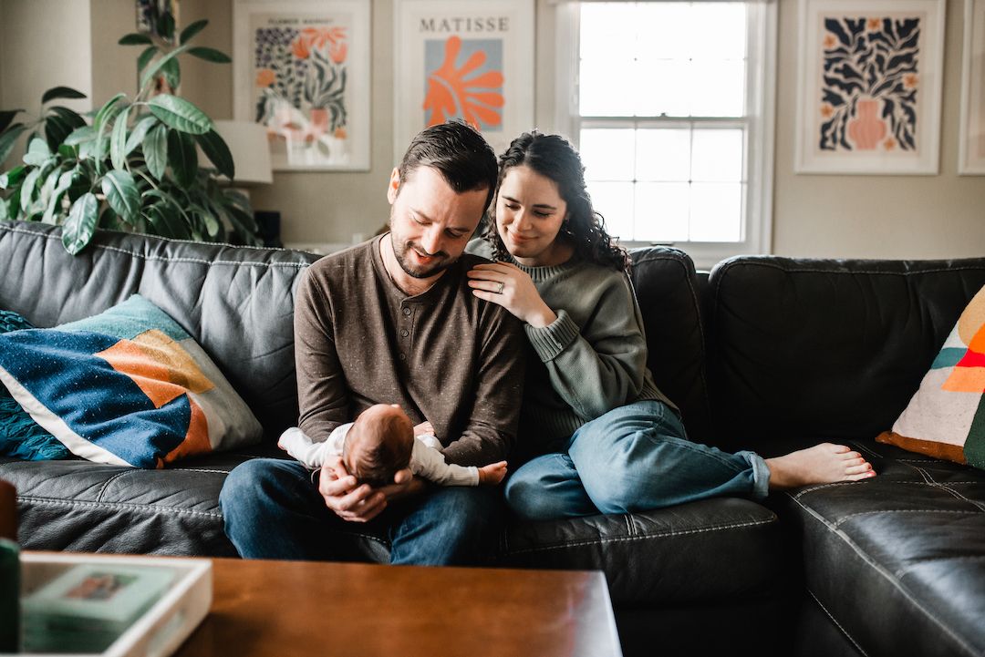 A couple sits on a leather sofa, tenderly cradling their newborn baby in a cozy, art-decorated living room.