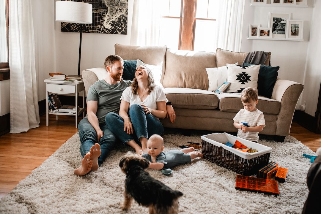 Happy family with baby, toddler, and dog relaxing together on living room floor.