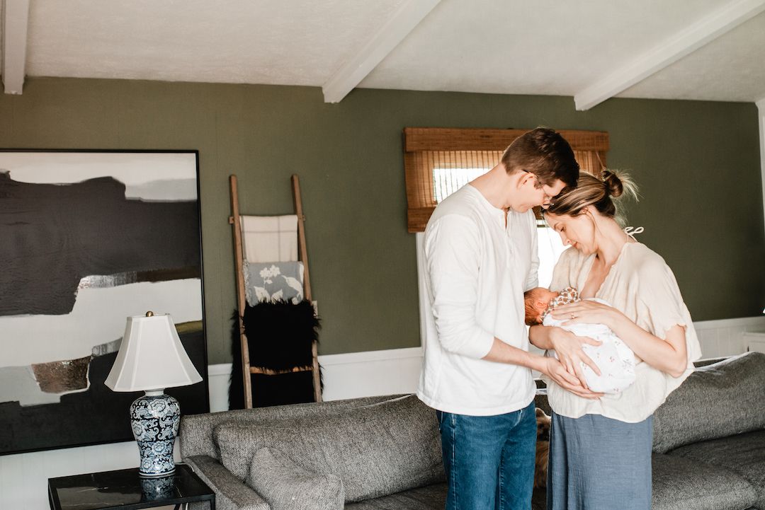 New parents stand together in a cozy living room, lovingly cradling their newborn baby in soft natural light.