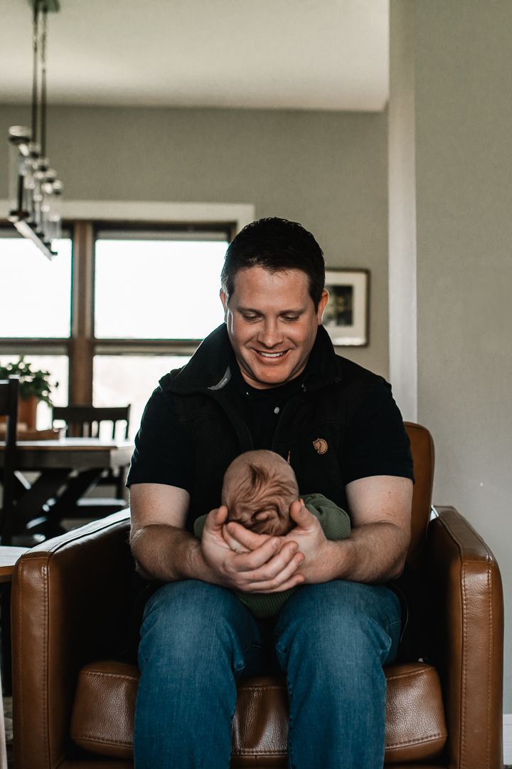 Smiling father sits in brown leather chair holding sleeping newborn baby in cozy home interior.