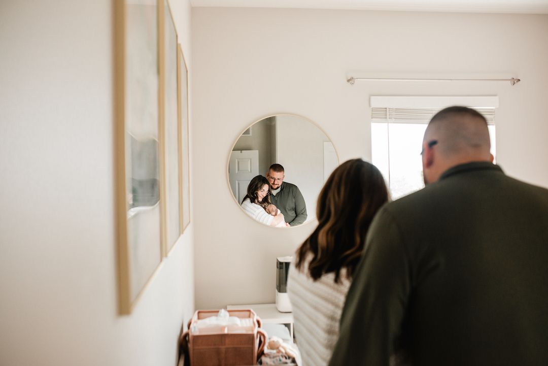 A couple admires their reflection holding a newborn in a round mirror in a bright modern home.