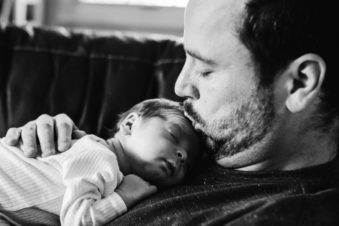 Father tenderly kissing his sleeping newborn baby in a black and white portrait photo.