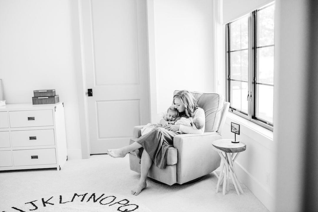 Black and white photo of a mother cuddling her child in a cozy nursery chair beside a bright window.