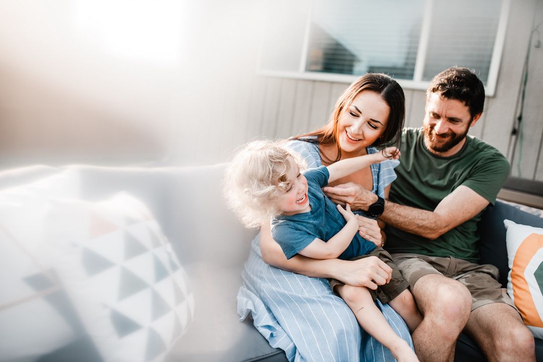 Smiling parents playfully tickling their young child on outdoor sofa.