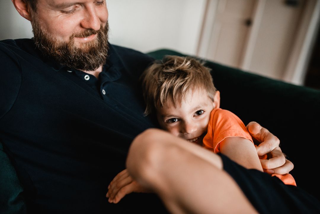 Bearded father cuddling toddler son on dark sofa in cozy home setting.