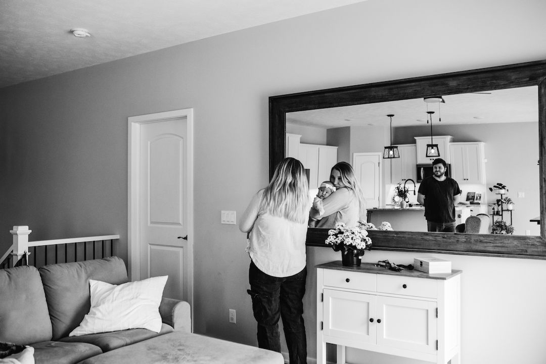 Black and white photo of a woman holding a baby near a mirror reflecting a man in a cozy home interior.
