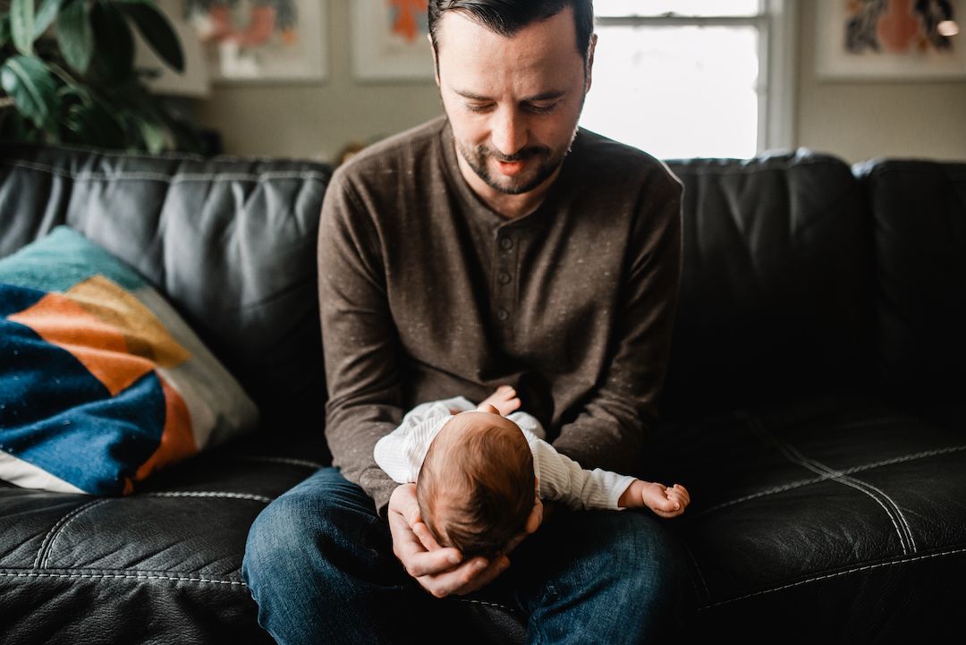 A father sits on a black leather sofa, gently cradling his newborn baby with a warm, loving smile.