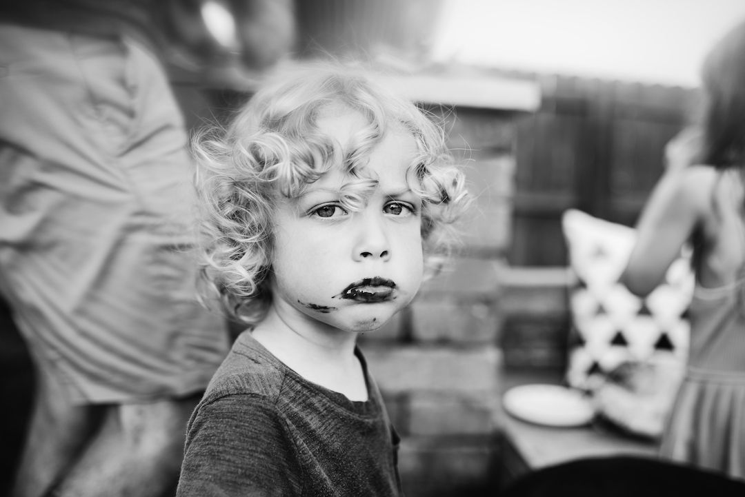 A curly-haired blonde child with a painted mouth looks seriously at the camera in black and white.