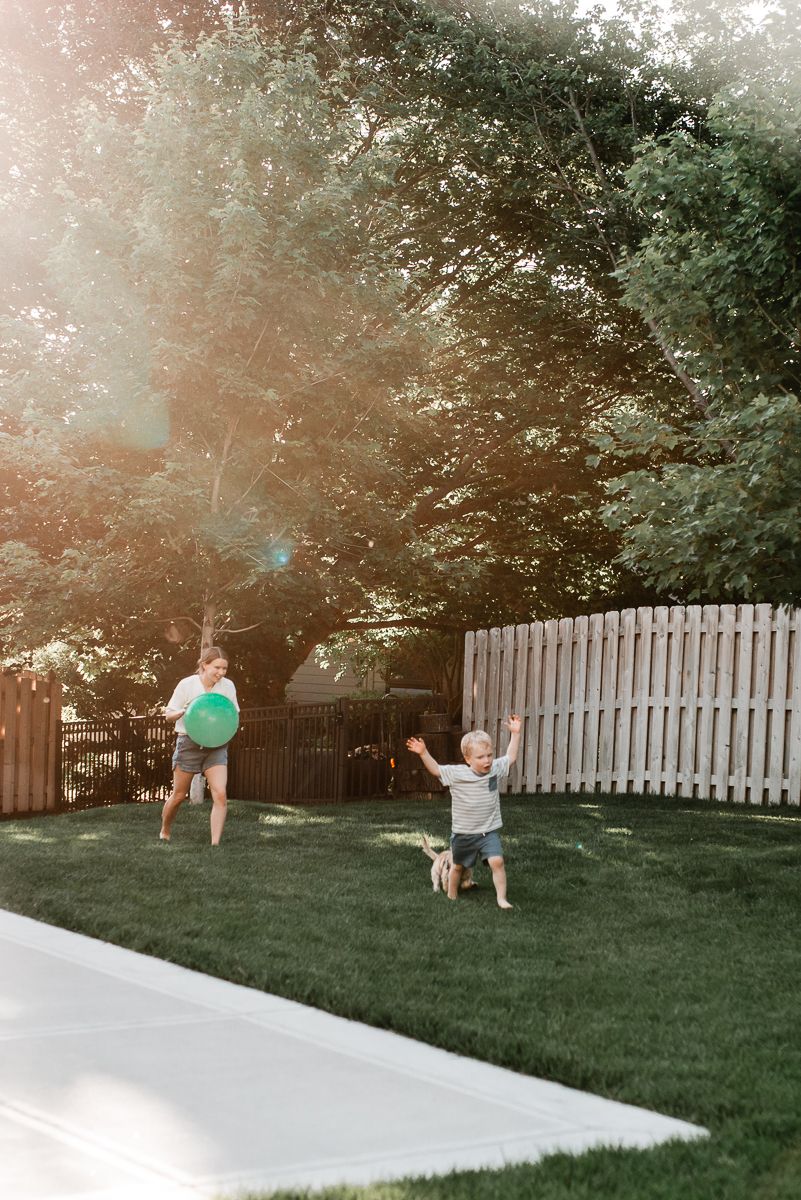 Two children playing in a sunny backyard, one holding a green ball while the other raises arms joyfully near a white fence.