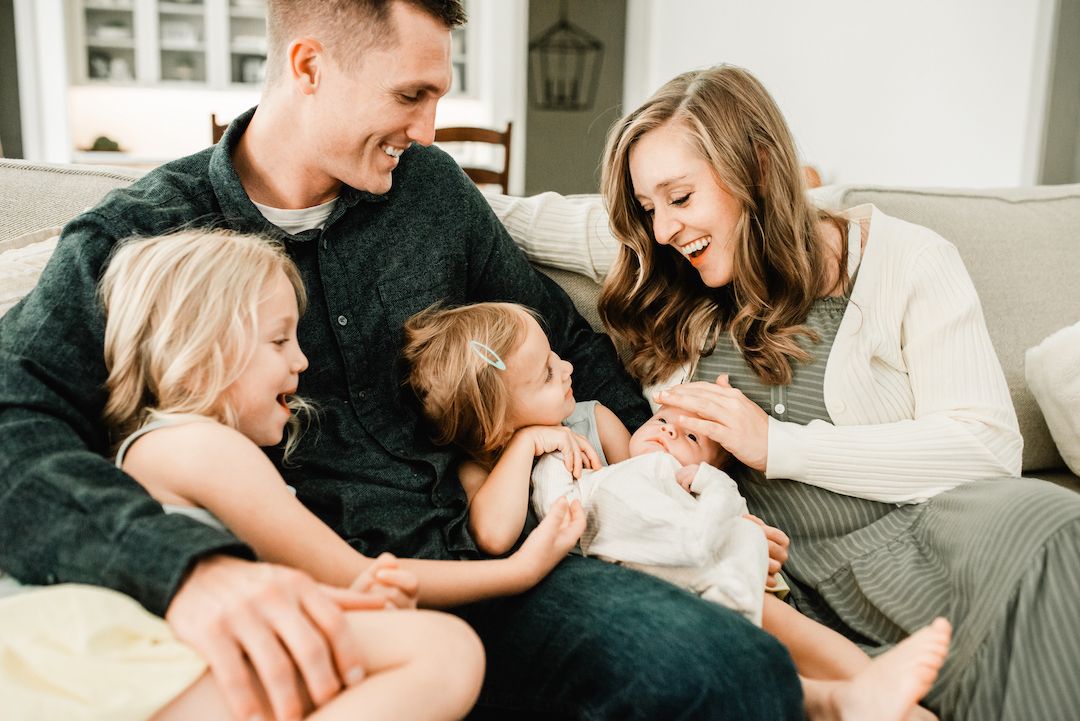 Happy family of four laughing together on a cozy living room couch, parents enjoying playful moment with two young children.