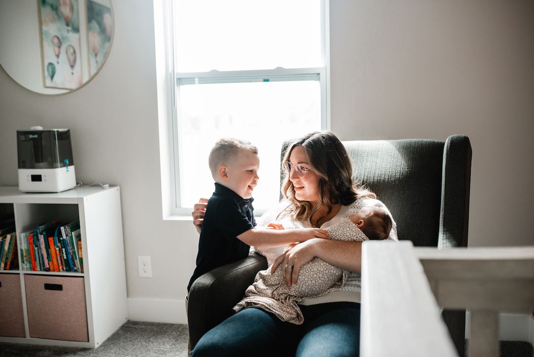 A mother sits in a gray chair holding a newborn while her young son leans in for a tender family moment.