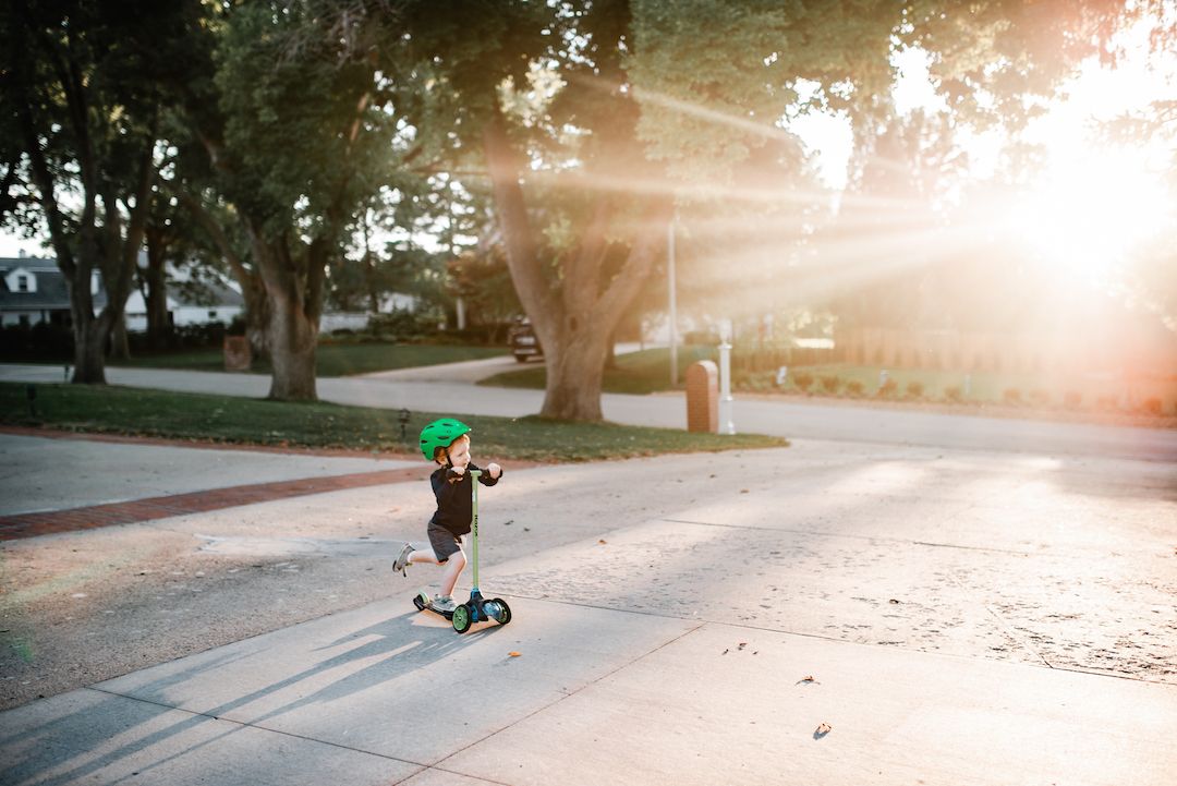 Child riding a scooter wearing a green helmet on a sunny suburban sidewalk with golden sunlight.