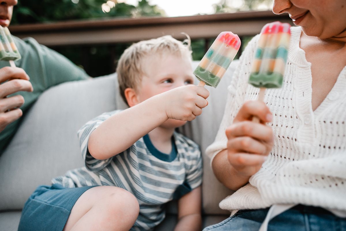 Family enjoying colorful popsicles outdoors on a couch, with a young blonde child holding a rainbow ice pop.