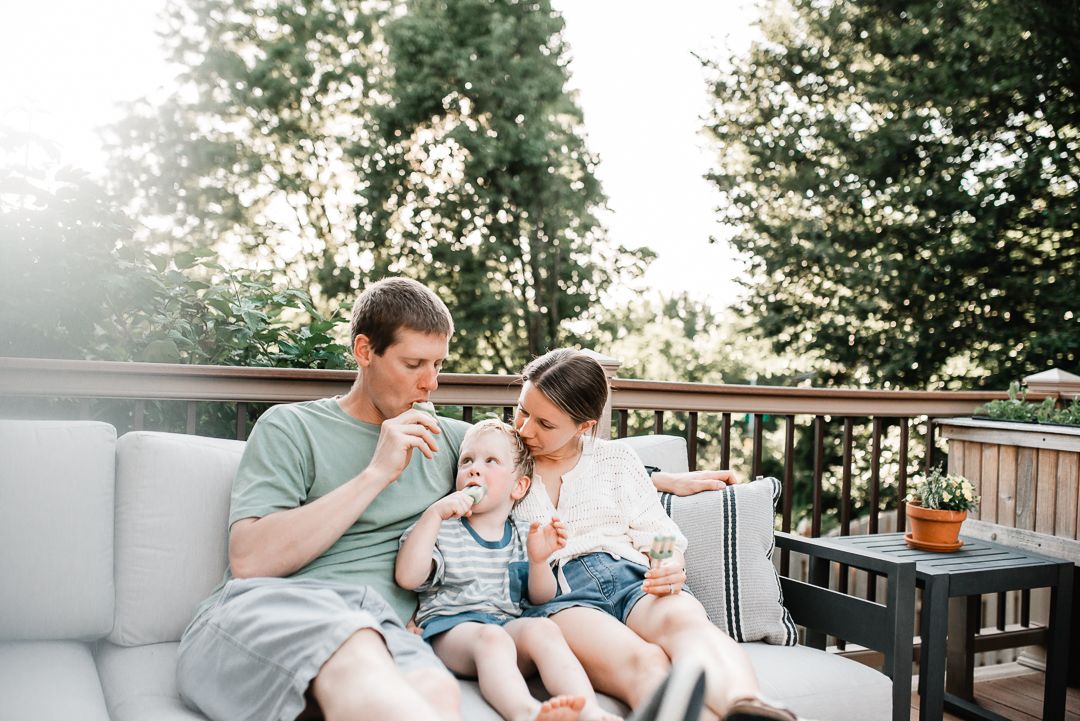 Family of three enjoying popsicles together on a cozy outdoor deck surrounded by lush green trees on a sunny day.