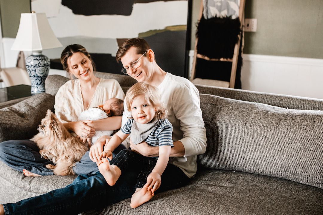 Smiling family with a toddler and newborn relaxing on a cozy gray sofa with a dog in a bright living room.