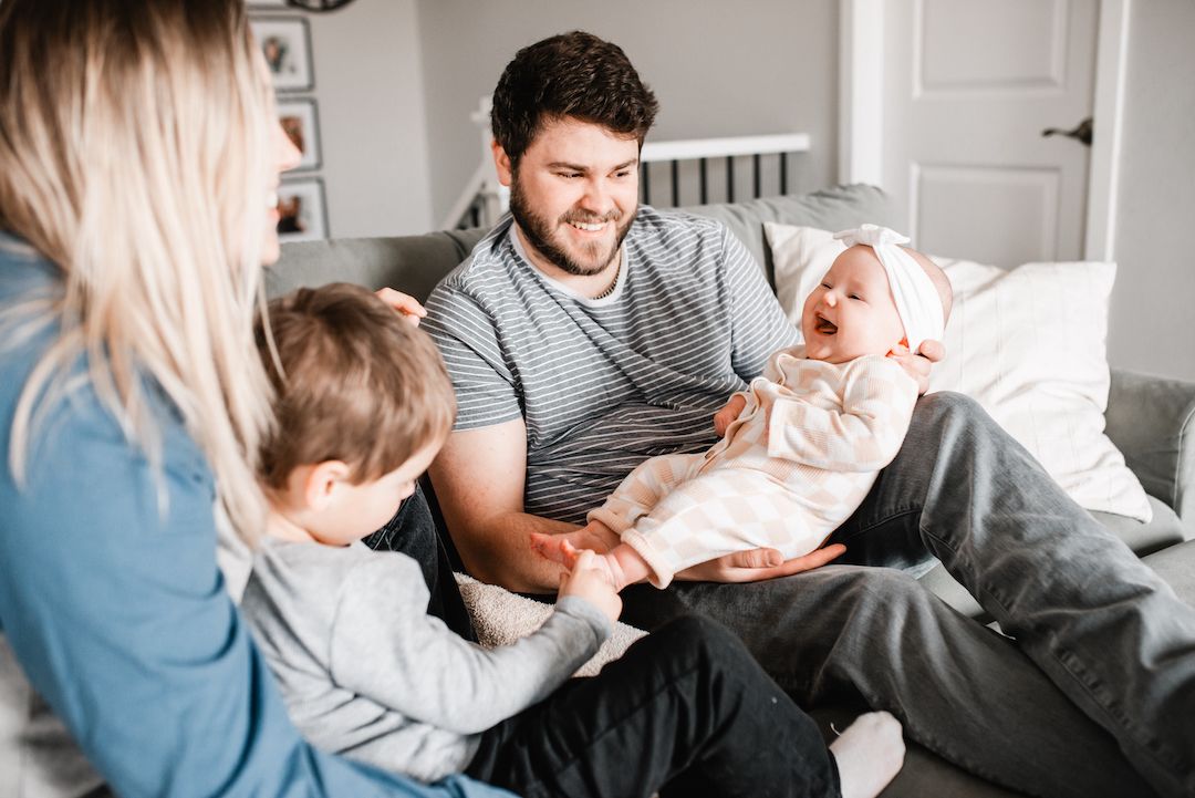 Happy family with toddler and laughing baby relaxing together on gray sofa at home.