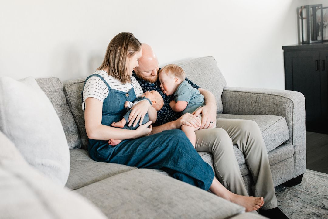 Family sitting on gray couch, mother holding newborn baby while toddler sits on father's lap in cozy living room.