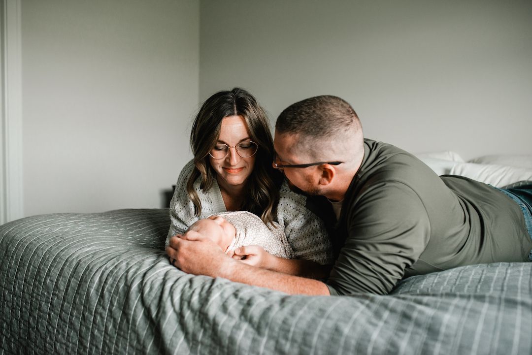 A loving couple lies on a bed, gazing adoringly at their newborn baby wrapped in a blanket.