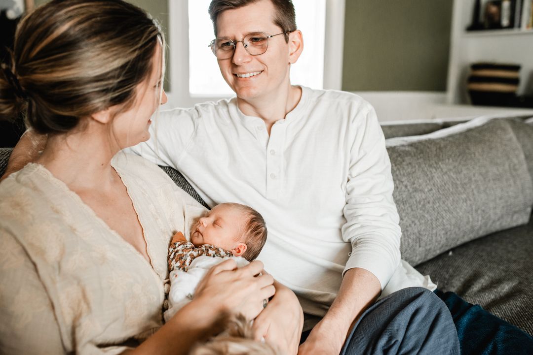 New parents smiling at newborn baby on couch at home.