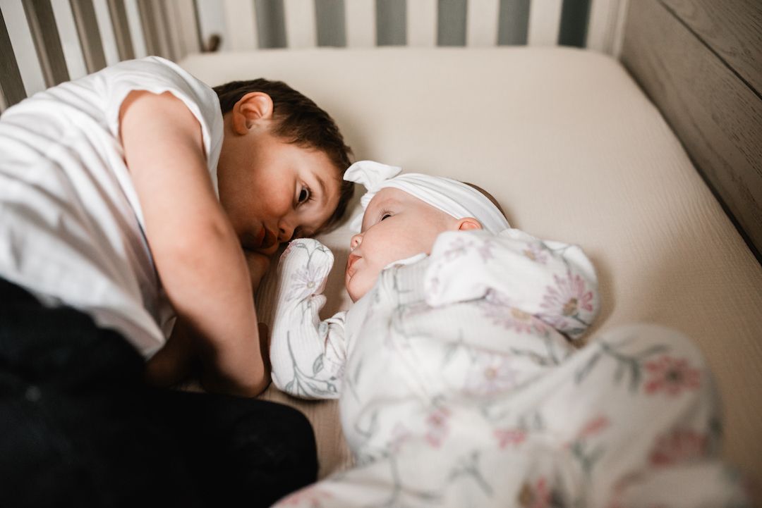 Older child leaning over crib to gently kiss newborn baby sibling wrapped in floral swaddle blanket.