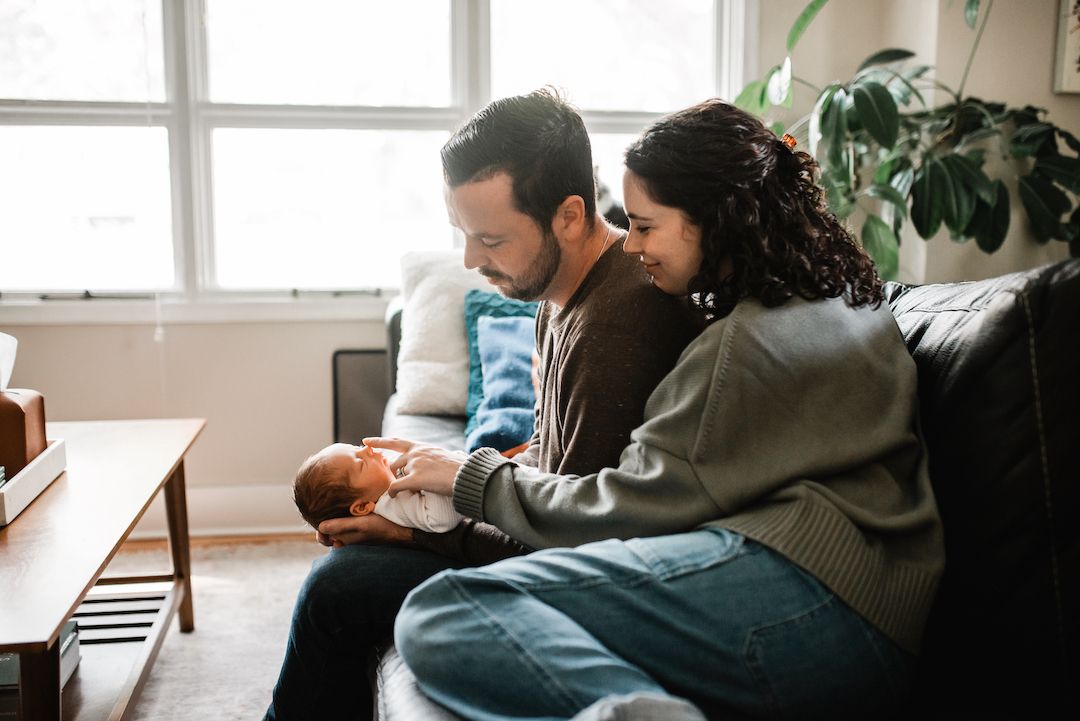 A couple sits on a couch together, lovingly gazing at their newborn baby in a bright living room.