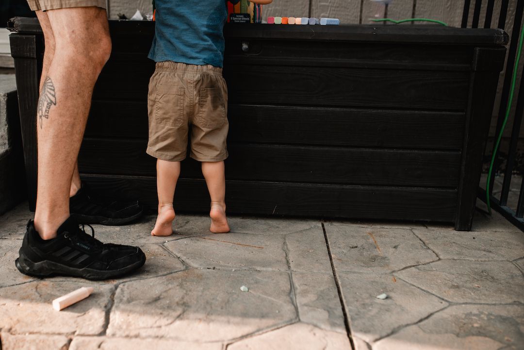 An adult with a tattoo stands beside a barefoot toddler on a stone patio.