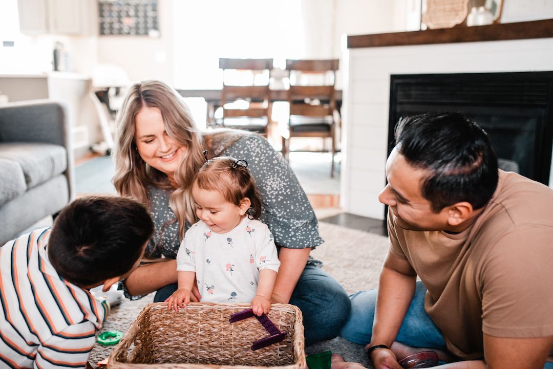 Happy family of four sitting on floor playing together near a fireplace.
