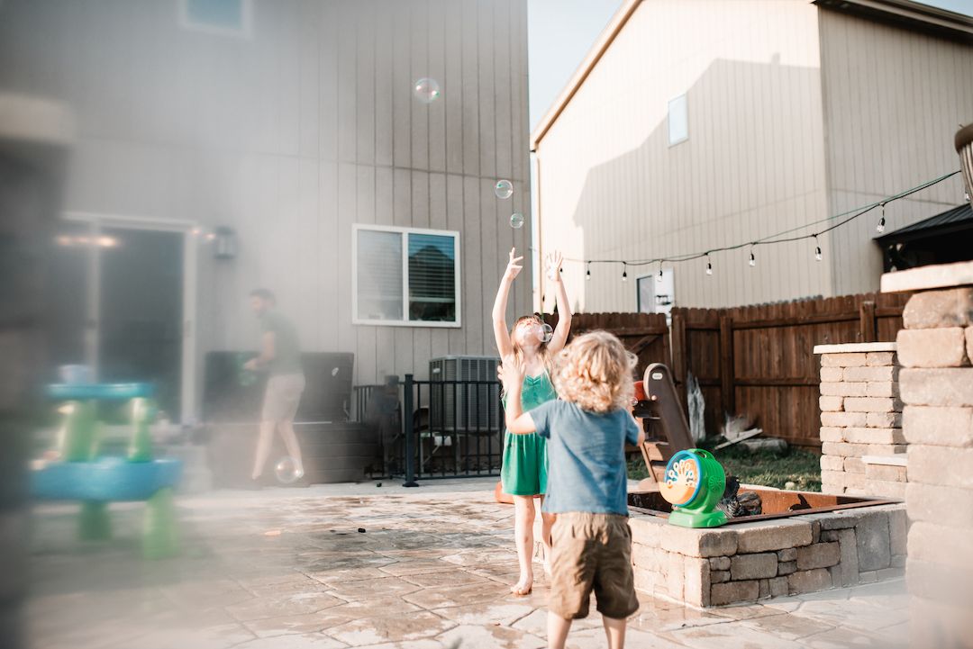 Two children chasing bubbles in backyard patio on a bright sunny day.