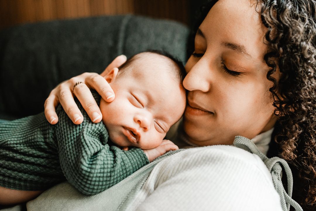 A mother tenderly holds her sleeping newborn baby dressed in green against her chest, sharing a peaceful bonding moment.