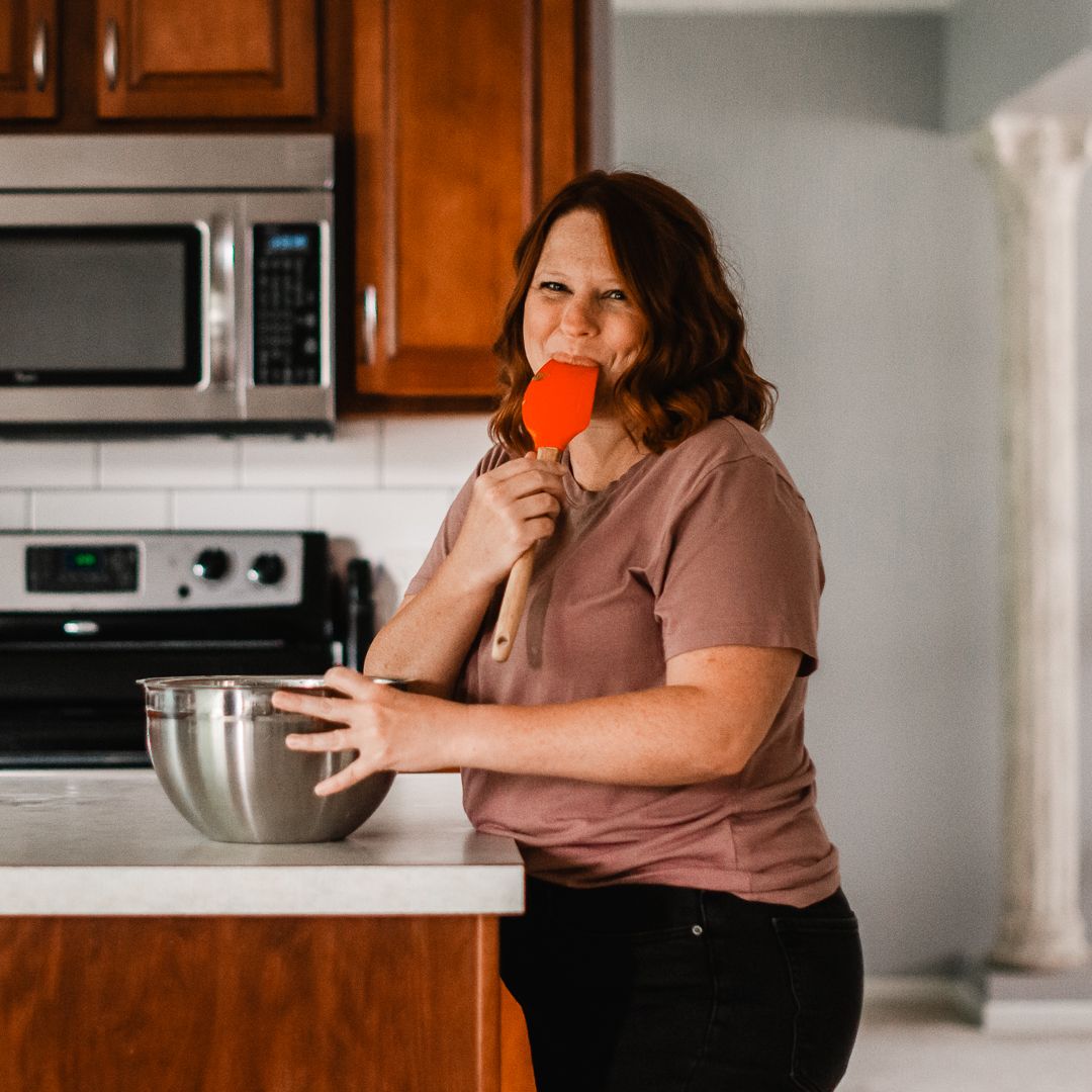 Woman licking a red spatula while standing at a kitchen counter with a mixing bowl, smiling playfully.