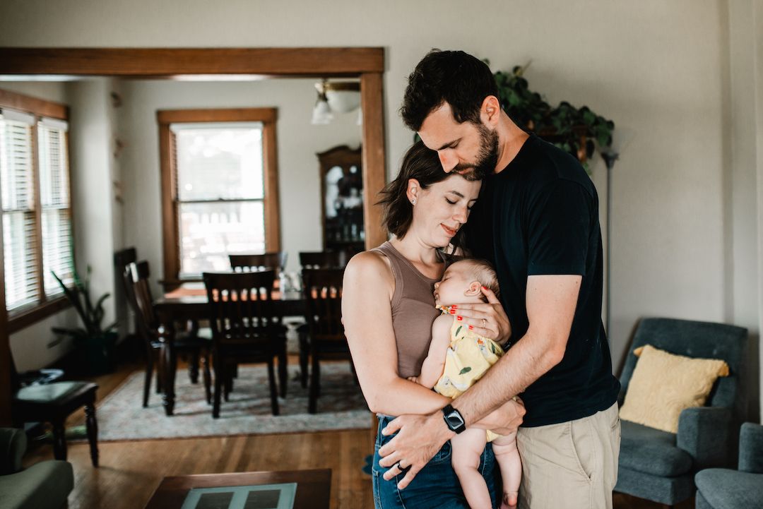 New parents standing indoors holding sleeping newborn baby wrapped in yellow blanket in bright home.