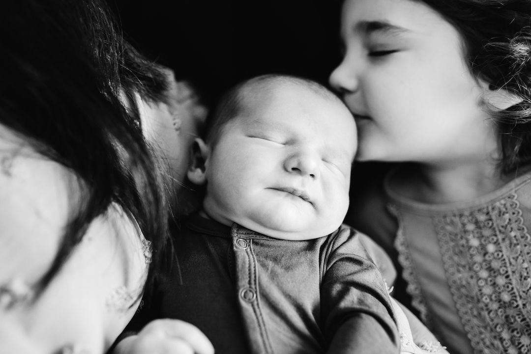 Black and white photo of a young girl kissing a sleeping newborn baby on the cheek.