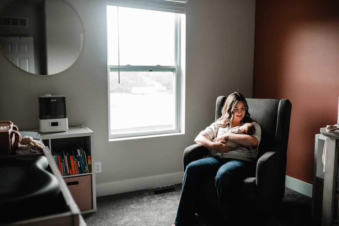 Mother sitting in gray armchair holding newborn baby near window in cozy nursery room.