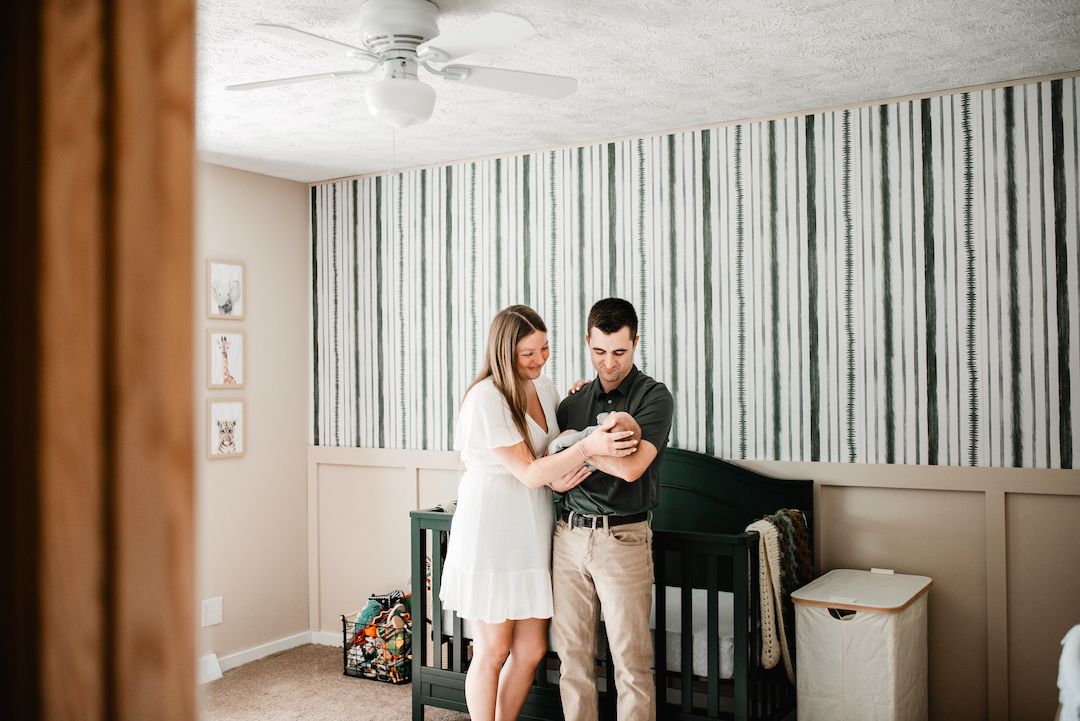 New parents admire their newborn baby together in a beautifully decorated nursery with striped wallpaper.