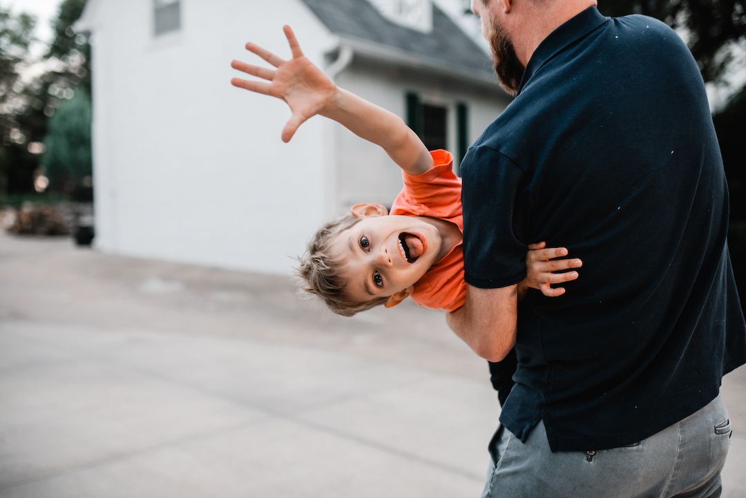 Father playfully holding laughing child upside down in front of home.