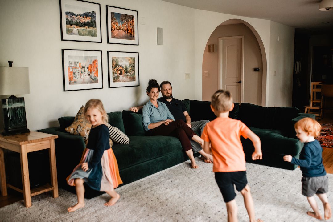 Two parents relax on a green sofa while their three young children play and dance in the living room.