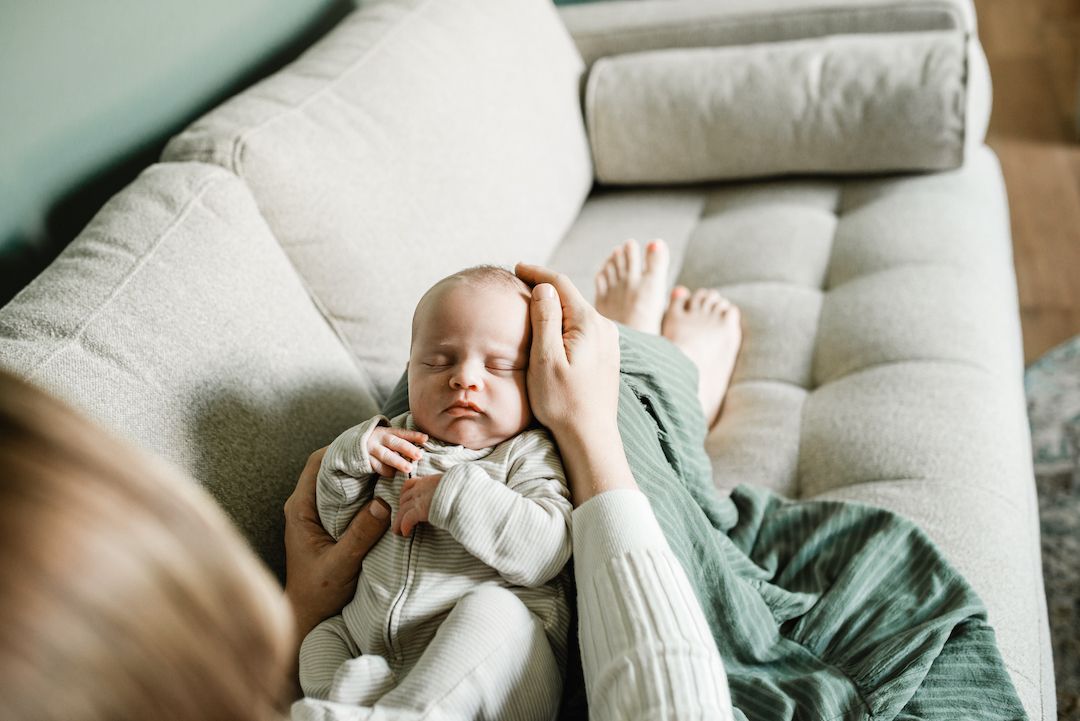 Mother gently cradling newborn baby on a light gray sofa, infant dressed in white onesie with green blanket nearby.