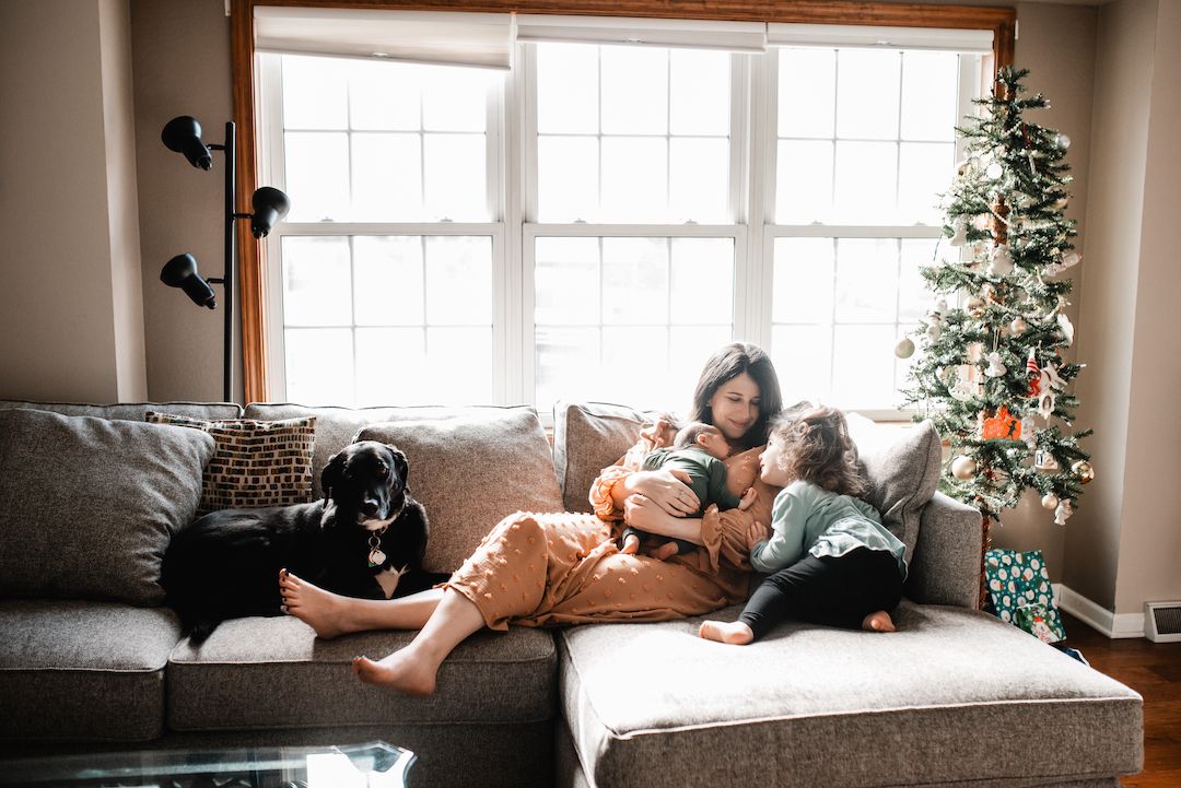 Family relaxing on gray sofa with black dog near Christmas tree in cozy living room.