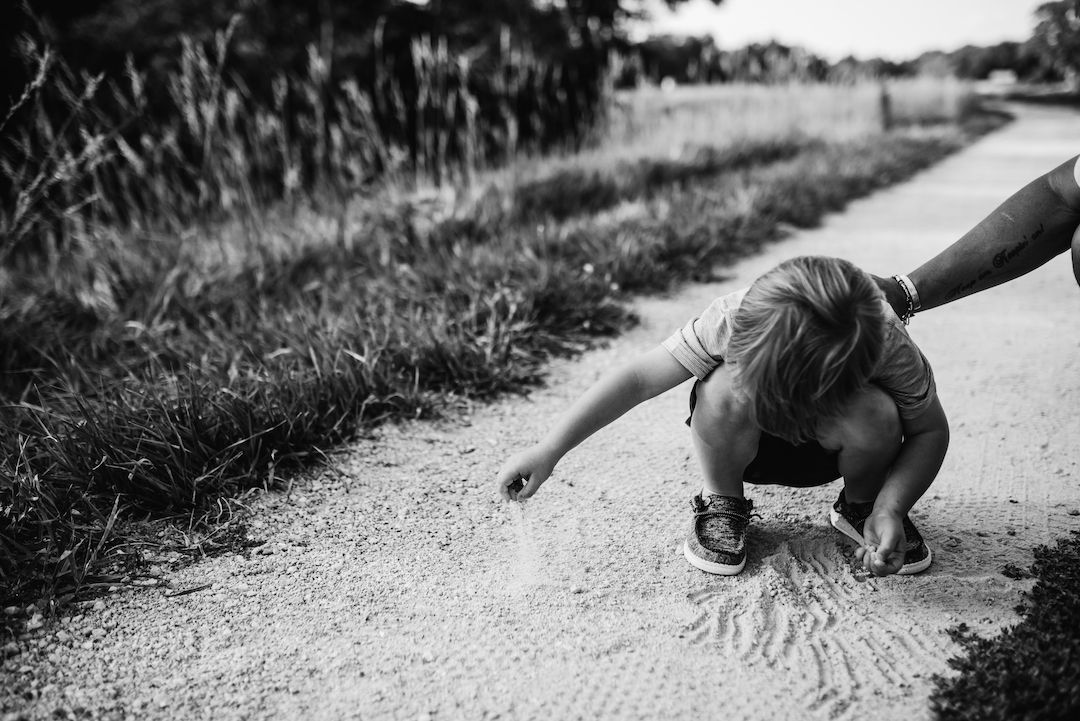Toddler crouching on a gravel path drawing in the dirt while an adult holds their hand.