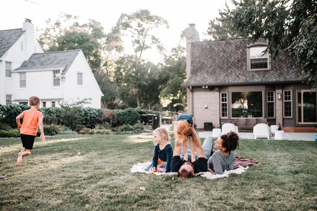 Family relaxing on blanket in backyard lawn at golden hour near charming white farmhouse.