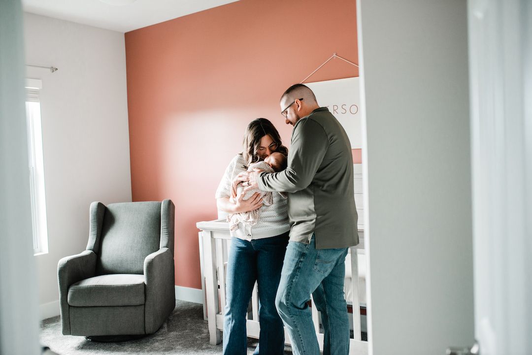 New parents admiring their newborn baby together in a nursery with a pink accent wall.
