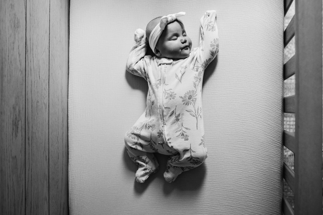 Black and white photo of a newborn baby lying in a crib wearing a floral onesie with arms raised.