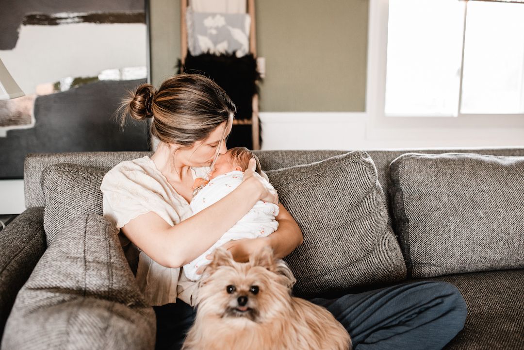A woman sits on a gray sofa cradling a newborn baby while a fluffy Pomeranian dog rests beside her.