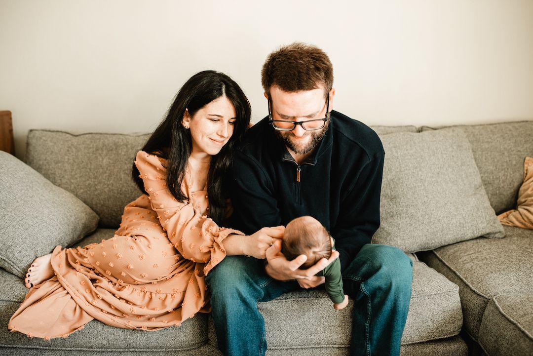 A couple sits on a gray couch, smiling as they look at their newborn baby together.