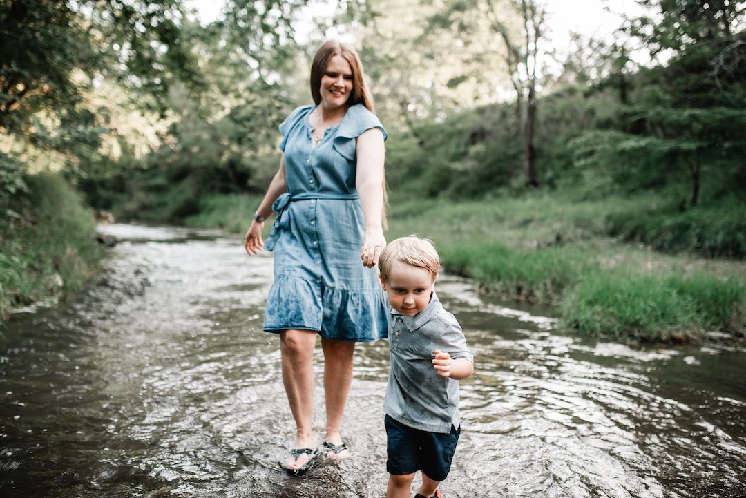 Mother and son walking through a shallow creek in a green forest setting.