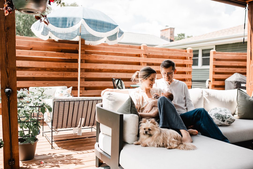 Couple relaxing on outdoor patio sofa with small dog, surrounded by wooden fence and lush greenery.