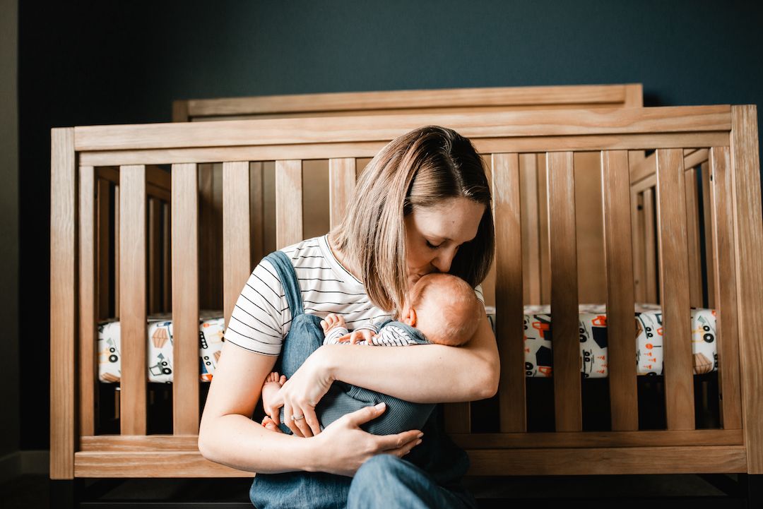Mother tenderly kissing her newborn baby while sitting in front of a wooden crib in a nursery.