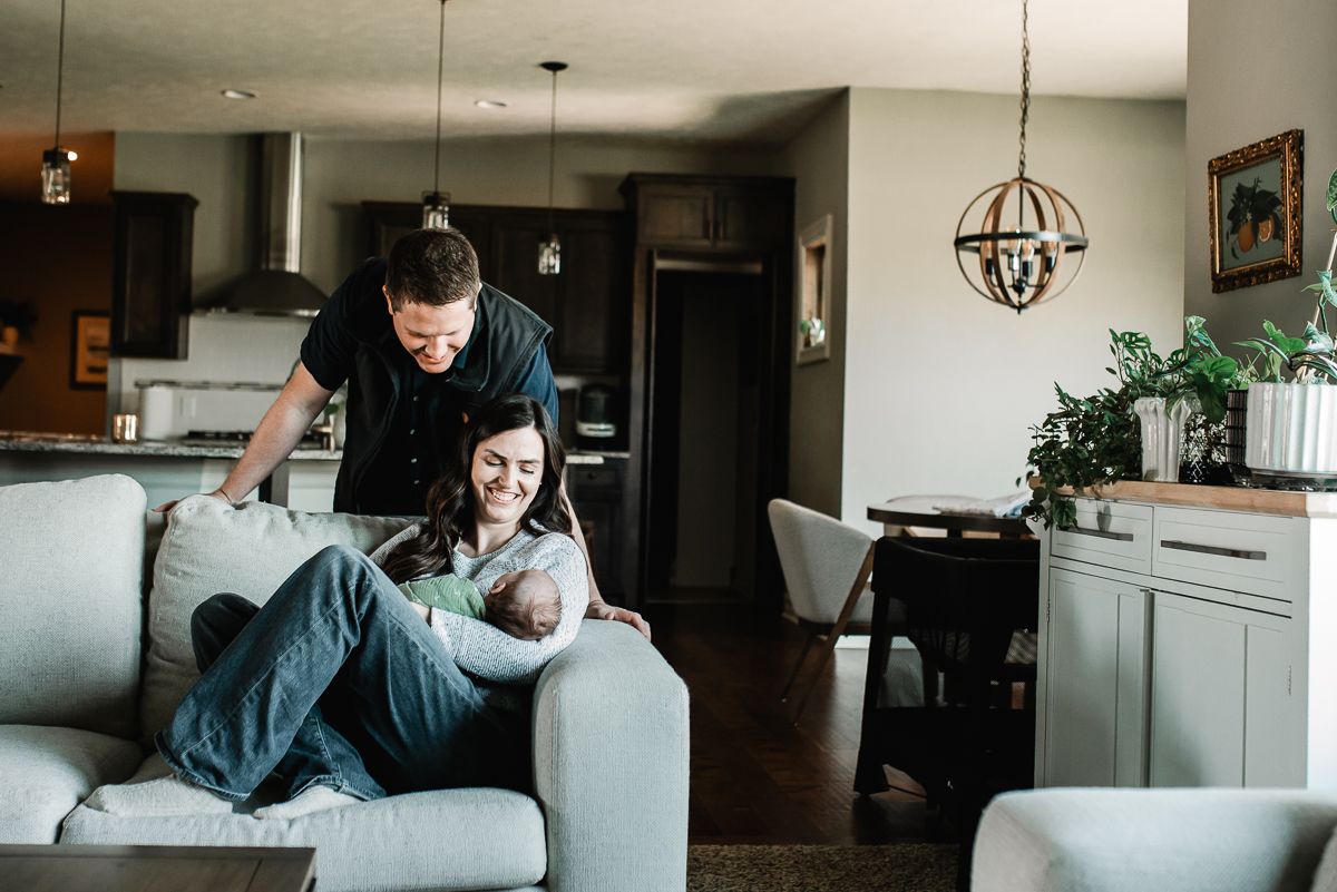 Couple relaxing on a gray sofa in a modern open-plan living room, man standing behind woman holding a newborn baby.