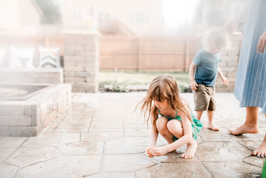 Young girl crouching on stone patio outdoors while child and adult stand nearby.