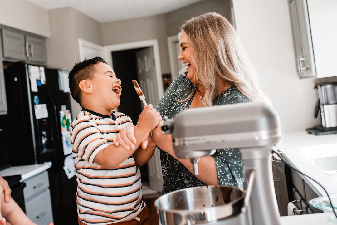 A woman and young boy laugh together while baking in a kitchen, sharing a fun moment with a mixer nearby.